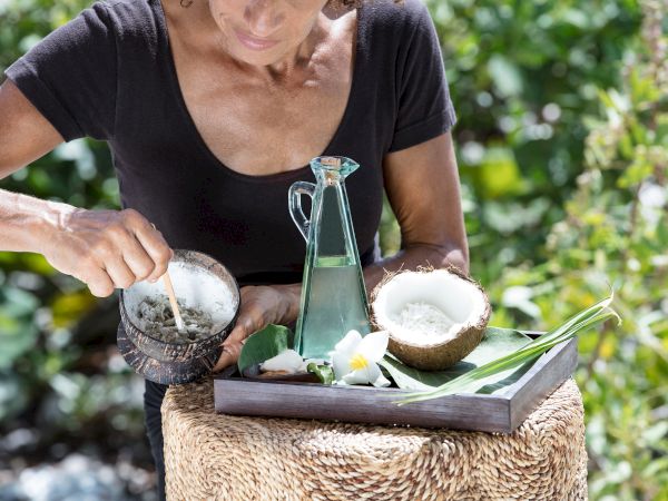 A woman pours sugar into two bowls on a tray with a glass bottle, lemons, and tropical leaves outdoors on a woven stool.