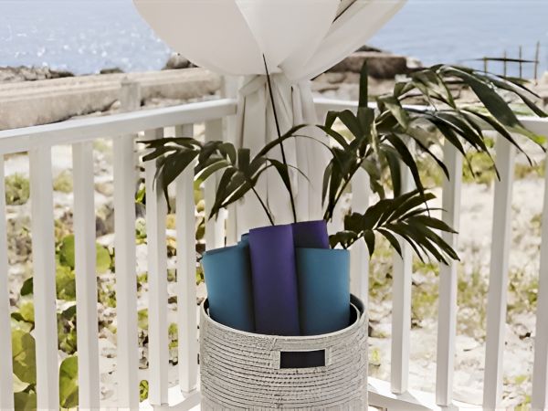 A white balcony with a woven planter holding three colored pots (blue, turquoise, purple) surrounding a leafy plant by the sea, under a canopy.