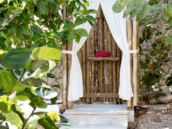 A small wooden doorway with white drapes tied back, nestled among green foliage and a red flower resting at the back.