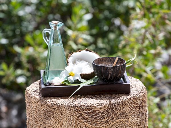A rustic scene: a glass bottle of water, a coconut, a wooden bowl, and a small tray with flowers, all on a woven pedestal outdoors.