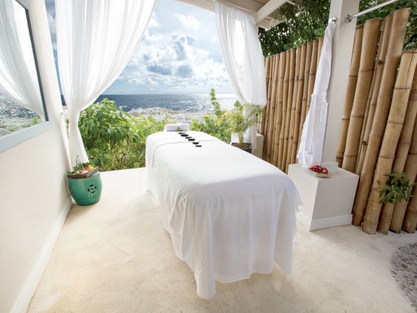 A serene outdoor spa setup with a white massage table under flowing curtains, overlooking a distant city view and lush greenery.