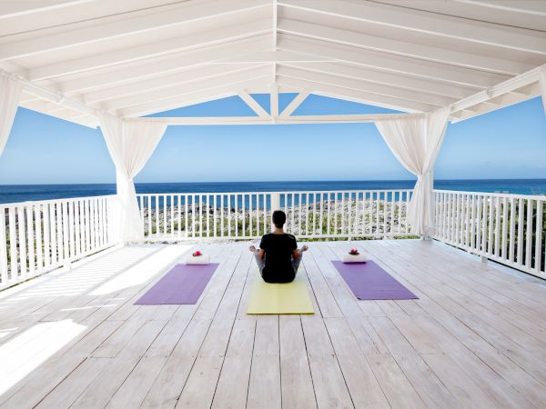 Three people practice yoga on mats inside a bright, open-air pavilion overlooking a blue ocean and clear sky, with white railings and curtains.