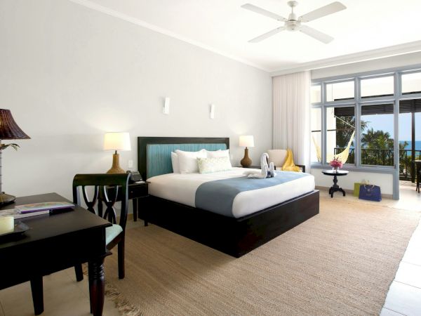A bright hotel room with a king bed, matching nightstands, lamps, and a desk; balcony doors open to a view outside.
