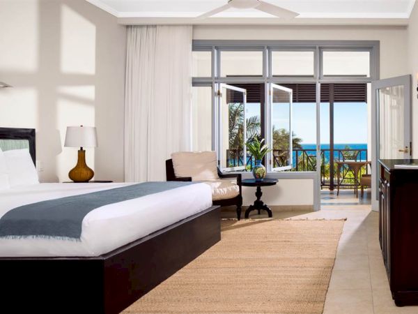 A bright hotel room with a large bed, a seating area, and a balcony overlooking the ocean through glass doors.