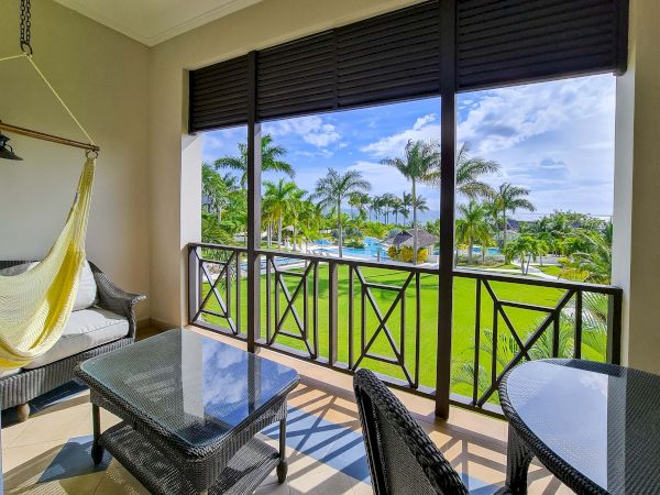 A balcony with wicker furniture overlooking a lush lawn, palm trees, and a sunny pool area beyond the railing, under a bright blue sky.