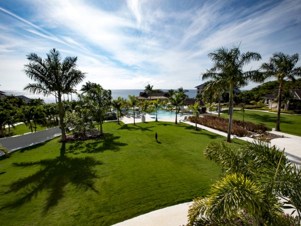 A sunny resort lawn with palm trees, a pool, pathways, and ocean view in the distance, inviting and tropical.