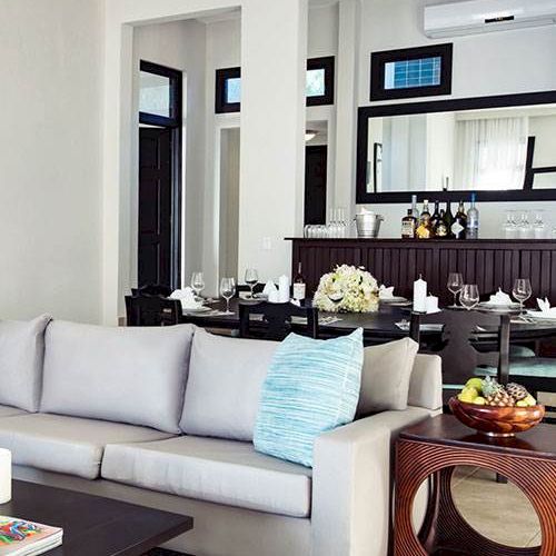 A modern, open living area with a light gray sofa, blue accent pillow, and a stylish dining space in the background. The room looks bright and elegant.