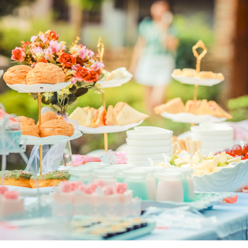 A colorful outdoor dessert table with cupcakes, pastries, fresh fruit, and flowers arranged on multi-tier stands and a bright blue cloth backdrop.