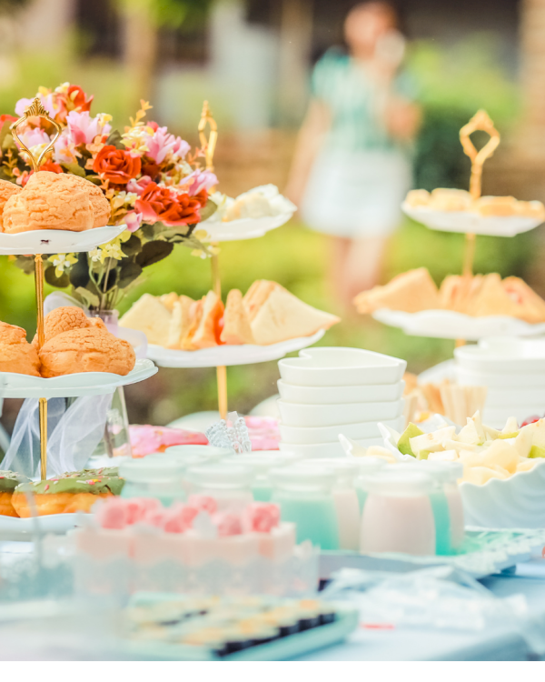 A colorful outdoor dessert table with cupcakes, pastries, fresh fruit, and flowers arranged on multi-tier stands and a bright blue cloth backdrop.