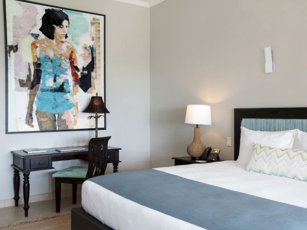 A modern hotel room with a large abstract artwork above a desk, a chair, and a bed featuring a blue-gray runner, soft lighting, and clean decor.