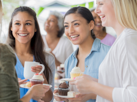 People socializing at a casual gathering, sharing cupcakes and drinks, smiling and chatting in a bright, friendly setting.
