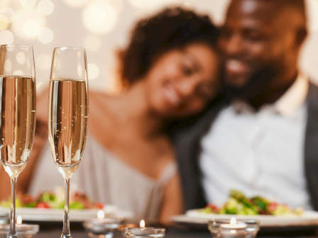 Two people at a romantic dinner with two champagne flutes in focus; couple in the background smiling over plates of food.