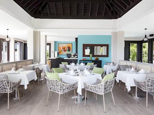 A bright, modern dining room with white tablecloths, wicker chairs, pastel accents, and large windows letting in natural light.