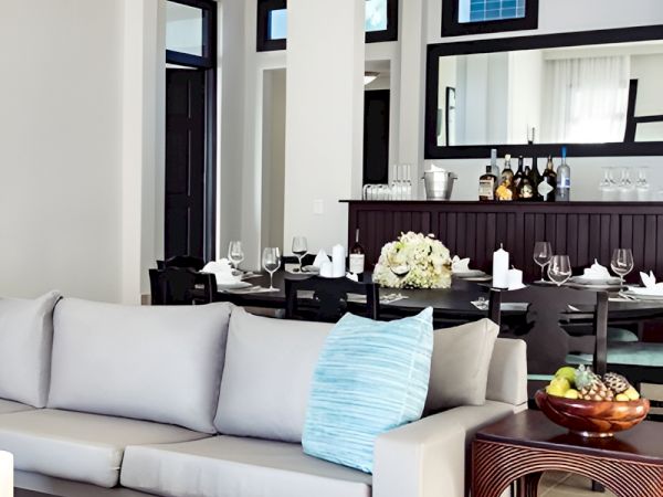 A modern, open-concept living and dining area with a light gray sofa, a teal-blue throw pillow, and a dark wood dining setup reflected in large mirrors.