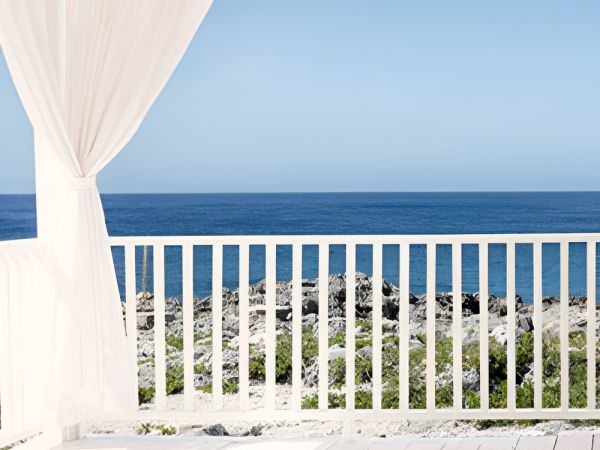 A bright seaside deck with white railings and sheer curtains, overlooking a calm blue ocean under a clear sky.