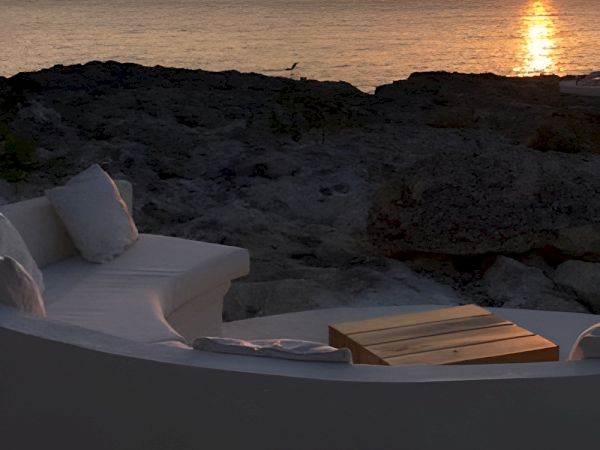 Sunset over a rocky beach with orange sky, calm sea, and a white outdoor sofa setup facing the water, cozy and serene.