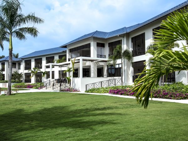 A modern white villa-style hotel with balconies, palm trees, and a green lawn under a blue sky, inviting outdoor relaxation.