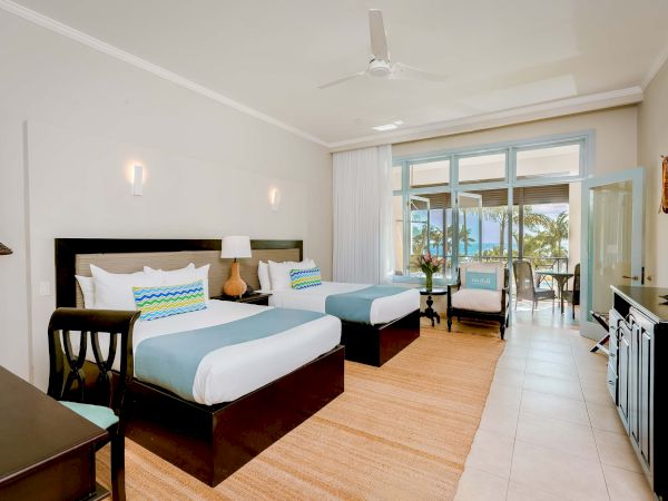 A bright hotel suite with two beds, a seating area, and a balcony; tropical decor, light wood floors, and glass doors to the outside.