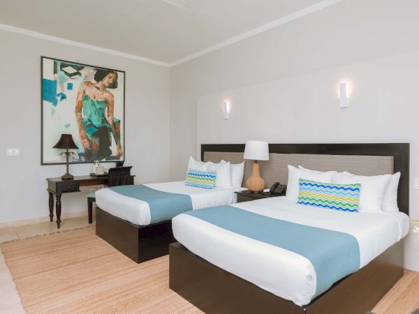 Two neatly made twin beds in a bright hotel room with a nightstand, lamps, art on the wall, and a beige rug.