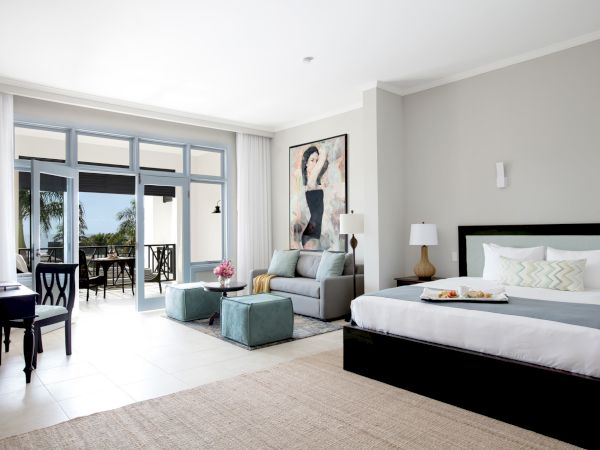A bright hotel-style suite with a king bed, a seating area, and sliding glass doors opening to a balcony, modern decor, and light neutral tones.