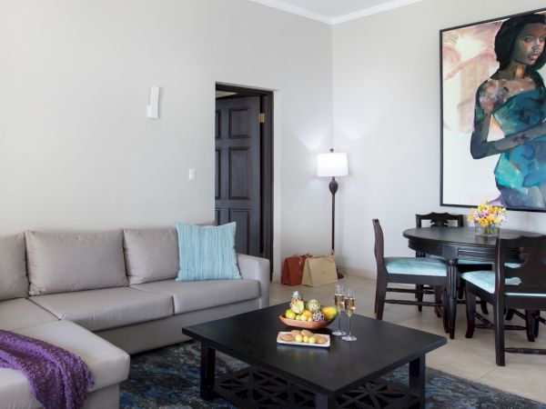 A modern living room with a beige sectional, blue pillows, a dark coffee table, a dining area with a chair set, a bright painting, and a floor lamp.