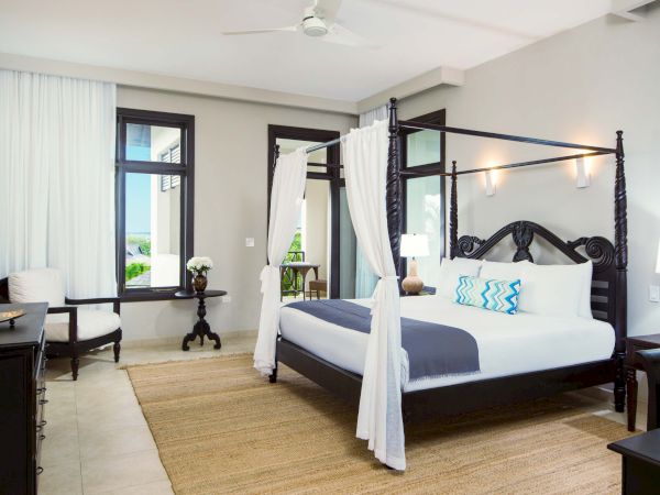 A luxurious four-poster bed with white drapes, dark wood frame, blue-and-white bedding, side tables, a rug, and a bright, airy hotel-style bedroom.