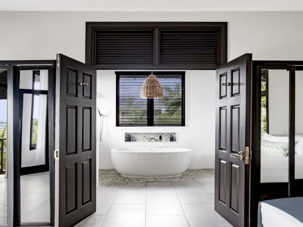 A bright, spa-like bathroom with a freestanding tub framed by dark double doors leading to a airy, modern suite.
