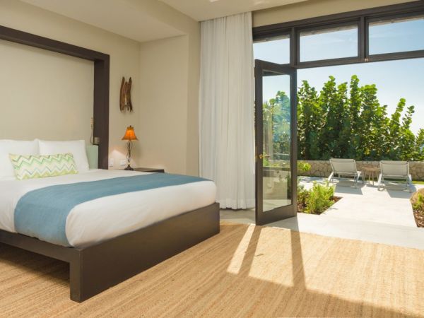 Modern hotel room with a large bed, dark wood furniture, and side lamps; sliding glass doors open to a sunny patio with lounge chairs.