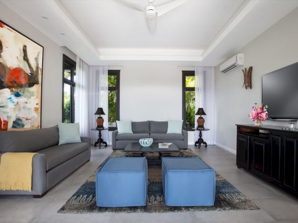 A bright, modern living room with a gray sectional, two blue ottomans, a large abstract painting, black cabinet, and condo-style decor.