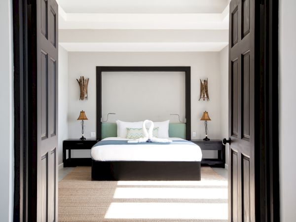 A bright, modern bedroom seen through dark double doors: a low bed with white and green bedding, two matching nightstands, and soft natural light.