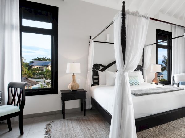 A bright, elegant hotel bedroom with a four-poster bed, white linens, sheer curtains, and soft lighting, plus a chair and lamps by the windows.