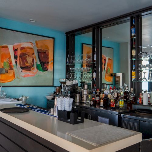 A modern bar with a sleek counter, glassware, and bottles behind, accented by turquoise walls and abstract art on the walls.