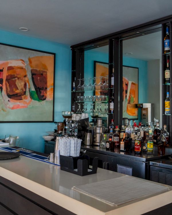 A modern bar with a sleek counter, glassware, and bottles behind, accented by turquoise walls and abstract art on the walls.
