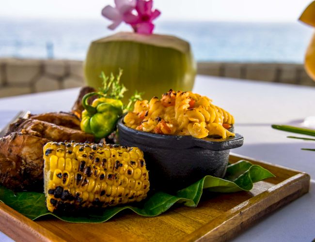 A tropical surfside meal: grilled meat, corn on the cob, and a skillet of vibrant fried rice or poke on a wooden board, with a coconut drink nearby.