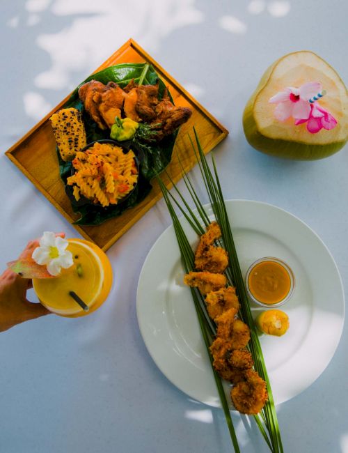 A vibrant spread of fried skewers and tempura on a plate with lime, dipping sauce, a tropical drink with a flower, and a carved lime garnish.