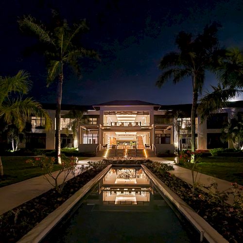 A modern house at dusk with warm lights, a reflecting pool, palm trees, and a tidy lawn&mdash;quiet, luxurious, and inviting.