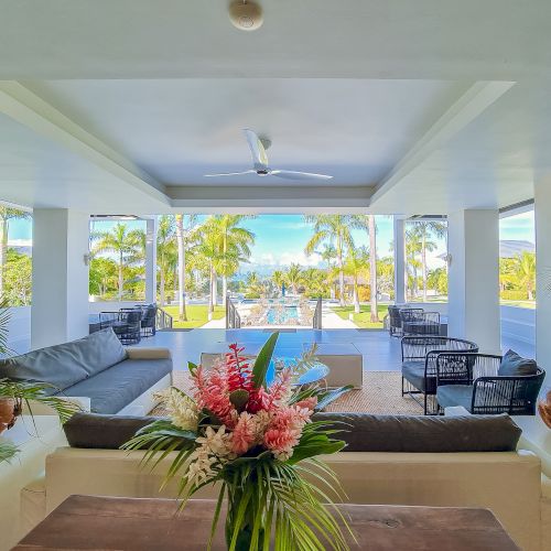 A bright, open living area with white walls, ceiling fan, and large windows showing palm trees and a pool; modern seating and tropical centerpiece.