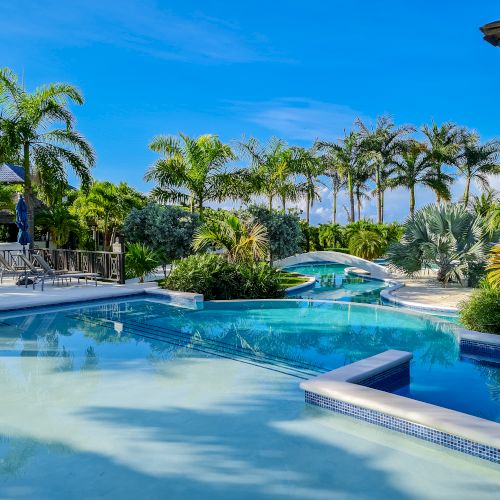A luxurious resort pool surrounded by palm trees, with clear blue water, sunlit lounge areas, and tropical landscaping.
