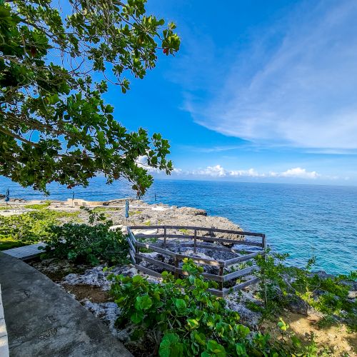 A sunny coastal view with a stone wall, lush greenery, and blue ocean meeting a bright sky, inviting a peaceful seaside stroll.