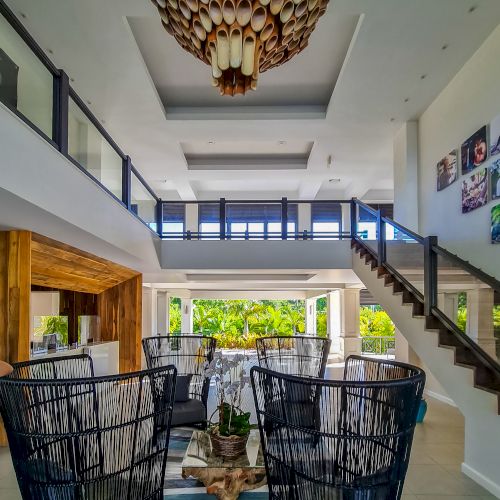 A bright, modern living/dining area with high ceilings, a woven pendant light, glass railing balcony, and stylish black chairs facing a coffee table.