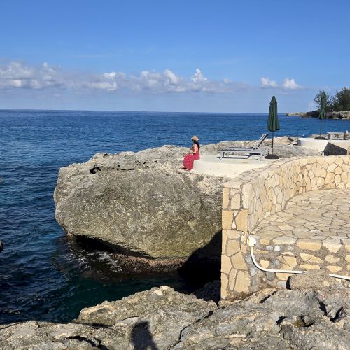 A person in a red outfit sits on a rocky ledge by the sea, with a stone walkway and clear blue water under a bright sky.