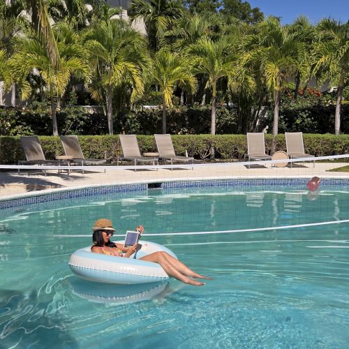 A person floats in a pool on an inflatable tube, reading a book, with palm trees and lounge chairs in the background.