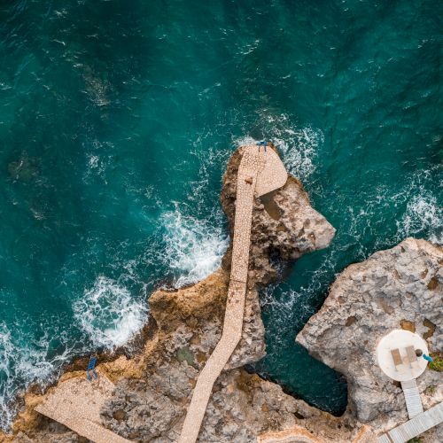 A rocky coastline with a concrete jetty extending into turquoise sea; two circular platforms perch at the edge, waves crashing below.