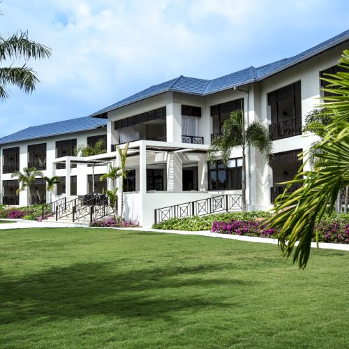 A large, elegant two-story white house with black shutters, a wraparound porch, well-kept lawn, and a sunny, shaded front yard.