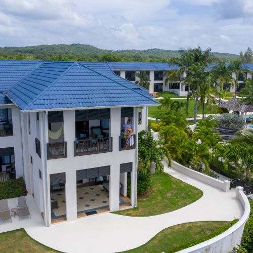 A two-story modern house with a dark roof, white and gray exterior, surrounded by a green lawn and palm trees, under a sunny sky.