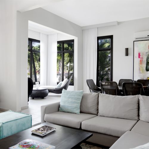 A bright, modern living room with a pale blue sofa, black coffee table, plants, and large windows bringing in natural light.