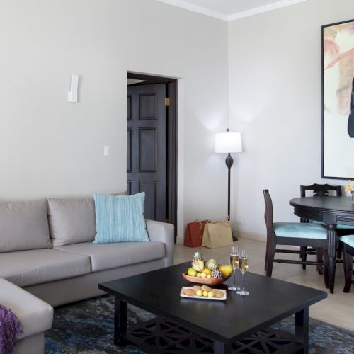 A bright, modern living room with a purple sofa, a cozy gray rug, wall shelves, and a person standing at a desk in the background, working.