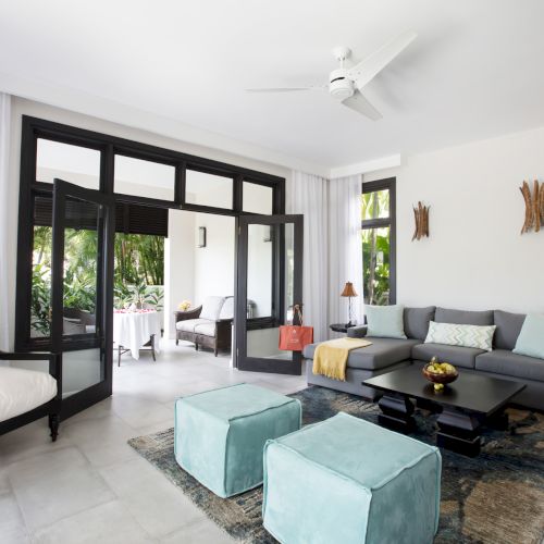 A bright, modern living room with large windows, a plaid sofa, glass coffee table, and a dining area in the background.