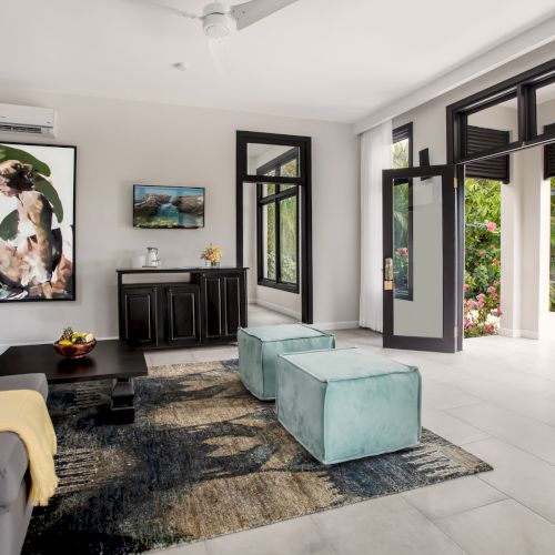 A bright, modern living room with a gray sofa, blue chairs, a white coffee table, and large windows letting in natural light.