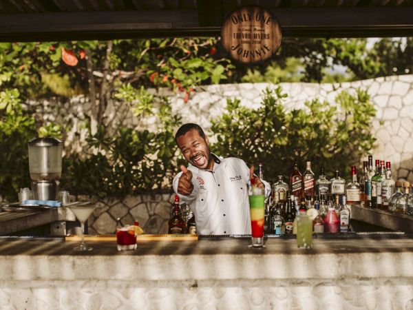 A bartender smiles and points at the camera behind a sunny outdoor bar with bottles and mixers, serving drinks with a friendly vibe.